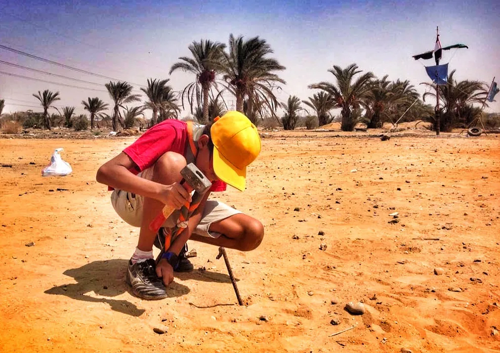 كشاف بقبعة صفراء يعمل على أرض صحراوية وأشجار النخيل خلفه
